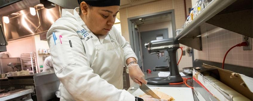 A student cutting a baked good with a knife in a kitchen.