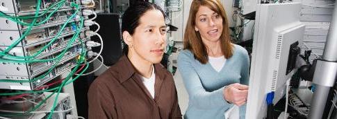 An instructor pointing out something on a monitor to a student in the foreground and a server rack in the background