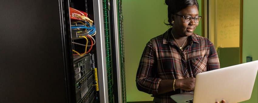 An Information Technology and Networking Student works in a server lab with a laptop making adjustments.